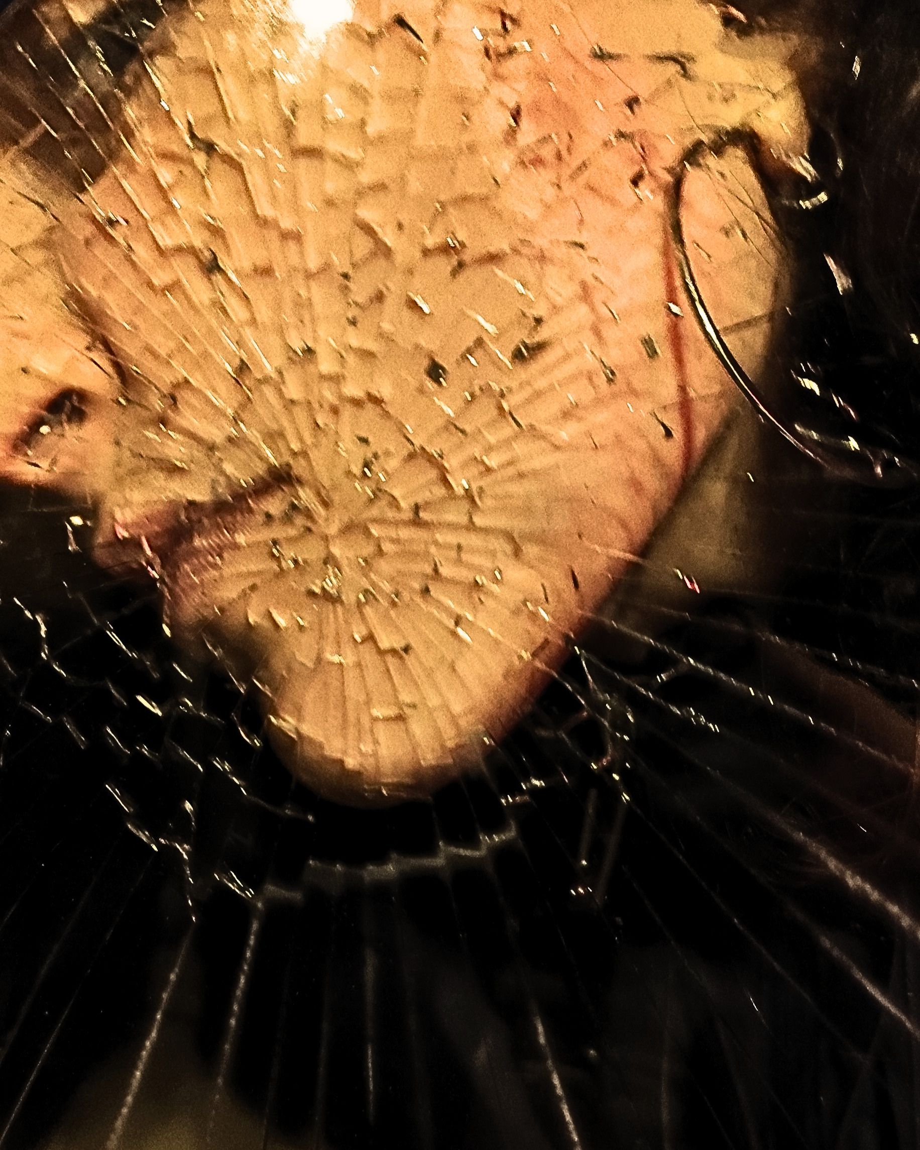 Nahaufnahme eines Gesichts hinter zerbrochenem Glas, das von Rissen bedeckt ist, die sich wie ein Spinnennetz über fast das gesamte Bild ausbreiten. Nur ein Auge ist sichtbar und blickt direkt in die Kamera; Wange, Nase und Lippen sind gegen die zerbrochene Oberfläche gedrückt, während an der Seite des Gesichts ein silberner Creolenohrring zu sehen ist.
