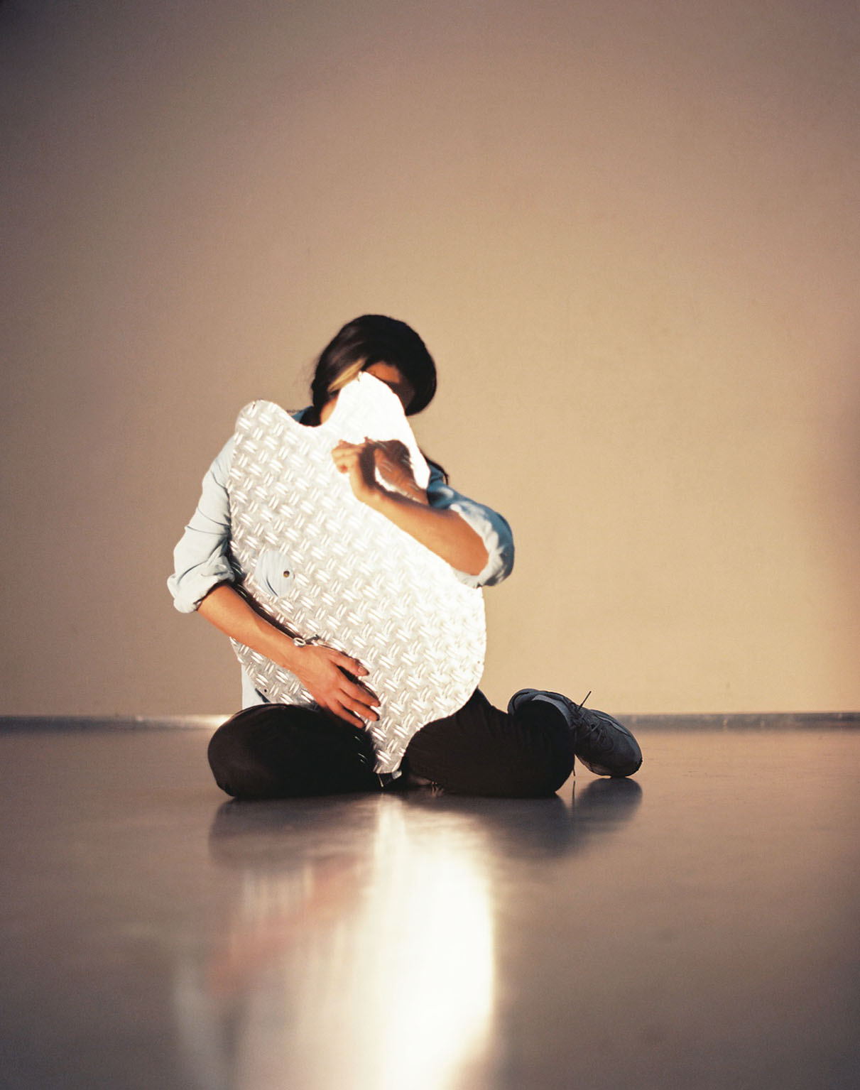 A person sits cross-legged on the floor, holding a large, reflective metal sheet that partially covers their face. The metal has a diamond pattern and catches the light, creating a soft glow against the otherwise neutral background. The person's posture appears calm and contemplative.