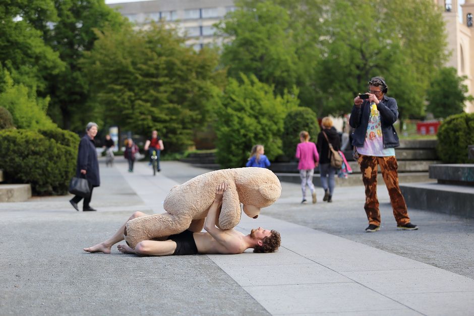 Eine nur mit Unterhose bekleidete Person liegt auf dem Rücken draußen auf einem Weg, auf ihr liegt ein Teddy, der fast so groß ist wie sie. Im Hintergrund sieht man vorbeilaufende Menschen, einer macht ein Foto von der liegenden Person.