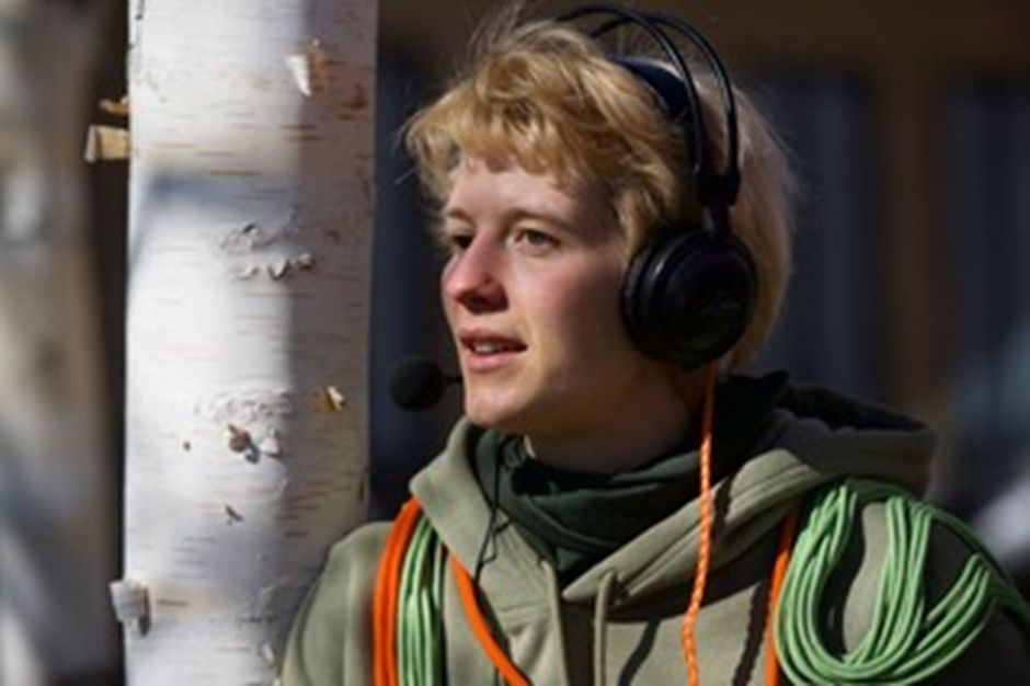 Abgebildet ist eine Draußenaufnahme von Fia Neises in der der Vorstellung „Ohren Sehen“. Ihr Oberkörper befindet sich in der Mitte des Bildes. Der Hintergrund zeigt den Stamm einer Birke. Ein Spiel aus Licht und Schatten erstreckt sich über die Fotografie. Fia Neises ist weiß, ca. 30 Jahre alt, hat kurze blond gefärbte Haare und grüne Augen, die in verschiedene Richtungen schauen. Ihr Gesicht wirkt offen und aufmerksam. Sie ist mit einem Kopfhörer und einem Headset ausgestattet. Sie trägt außerdem einen Olivfarbenen Kaputzenpullover, der mit grünen und orangenen Schnüren überwachsen ist.