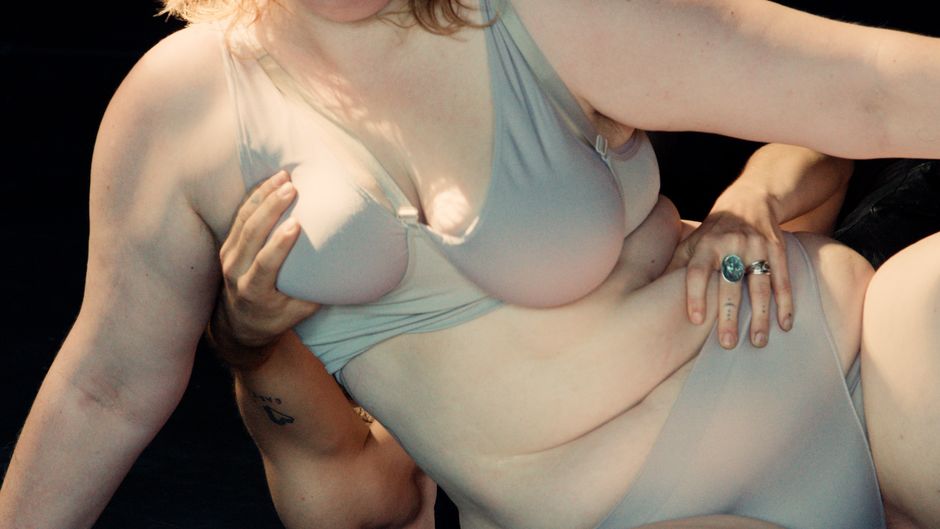 Eine Person mit hellem BH und Slip liegt halb seitlich. Eine Hand berührt den Bauch und eine andere Hand berührt die Brust der Person. Der Körper zeigt weiches Fett und Hautfalten, nah und intim fotografiert.