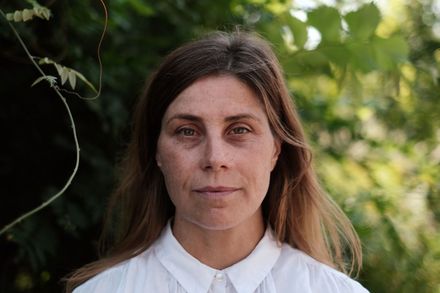 Ein Porträt der Choreografin Juli Reinartz. Sie steht vor einer grünen Pflanzenlandschaft. Sie hat lange dunkelblonde Haare und trägt eine weiße Bluse.