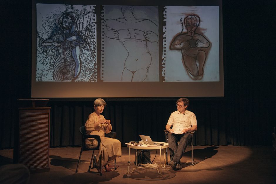 Zwei Personen sitzen an einem Tisch. Im Hintergrund hängt eine Leinwand, an die drei Zeichnungen von Körpern projiziert sind.