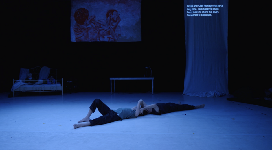 The picture shows a still from the performance EXTRA SET on the stage of HAU2, where the two dancers Márcio and Sindri lie on their backs, their heads touching and holding each other. The light is blue and dark. In the background hangs a painting depicting Márcio and Sindri. On the right side there is a white curtain with surtitles running across it.