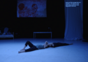 The picture shows a still from the performance EXTRA SET on the stage of HAU2, where the two dancers Márcio and Sindri lie on their backs, their heads touching and holding each other. The light is blue and dark. In the background hangs a painting depicting Márcio and Sindri. On the right side there is a white curtain with surtitles running across it.