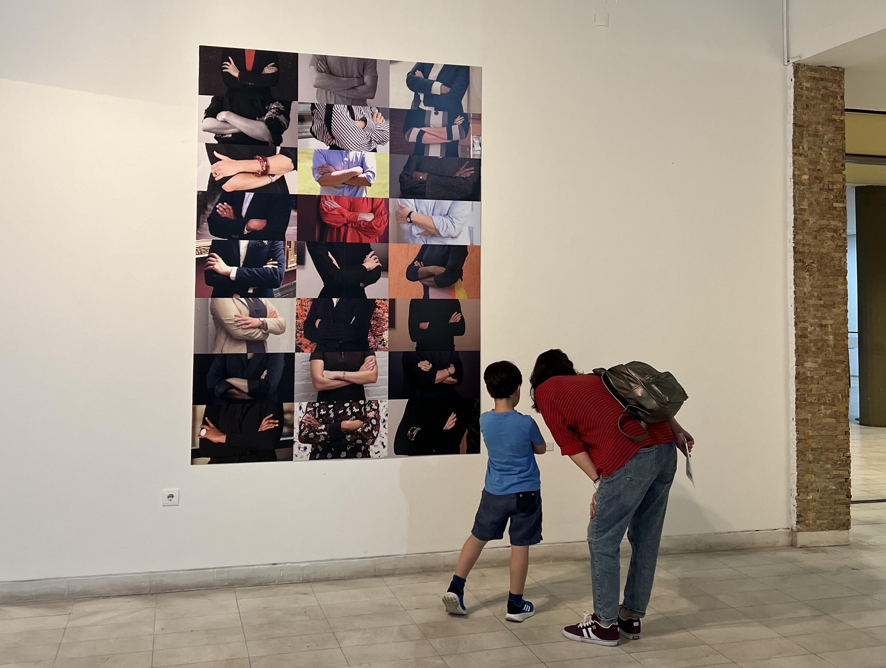 Eine erwachsene Person steht mit einem Kind vor einer Wand, an der eine Collage aus 24 Fotografien von verschränkten Armen hängt.